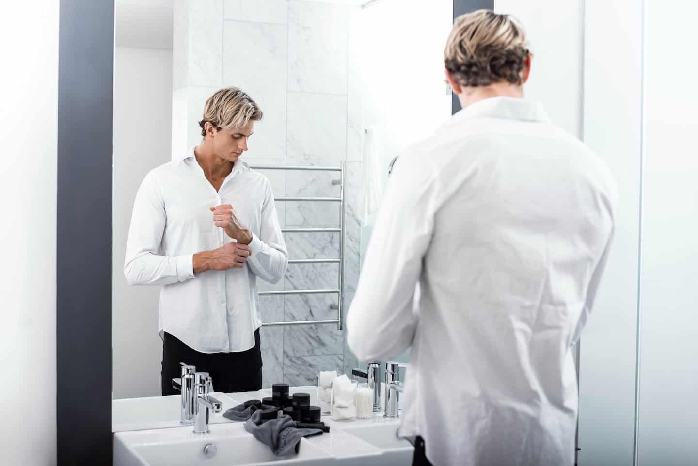 Man standing in front of mirror in white shirt buttoning cuffs