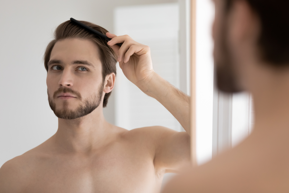 Handsome shirtless bearded 30s guy standing in bathroom after shower looks in mirror holding wooden comb, brushing his healthy hair. Ad of male haircare cosmetics products, beauty treatment concept