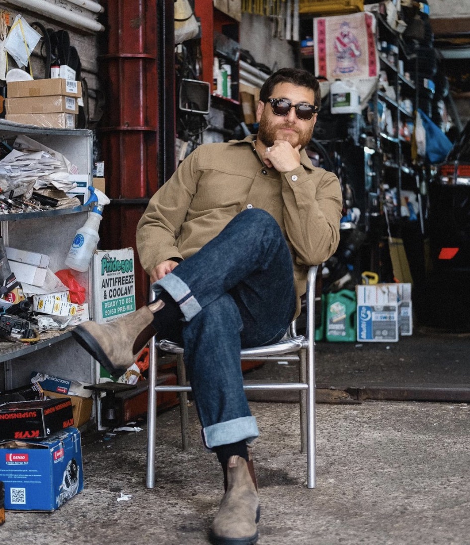 Man sitting in folding chair wearing cuffed jeans, sunglasses and jacket