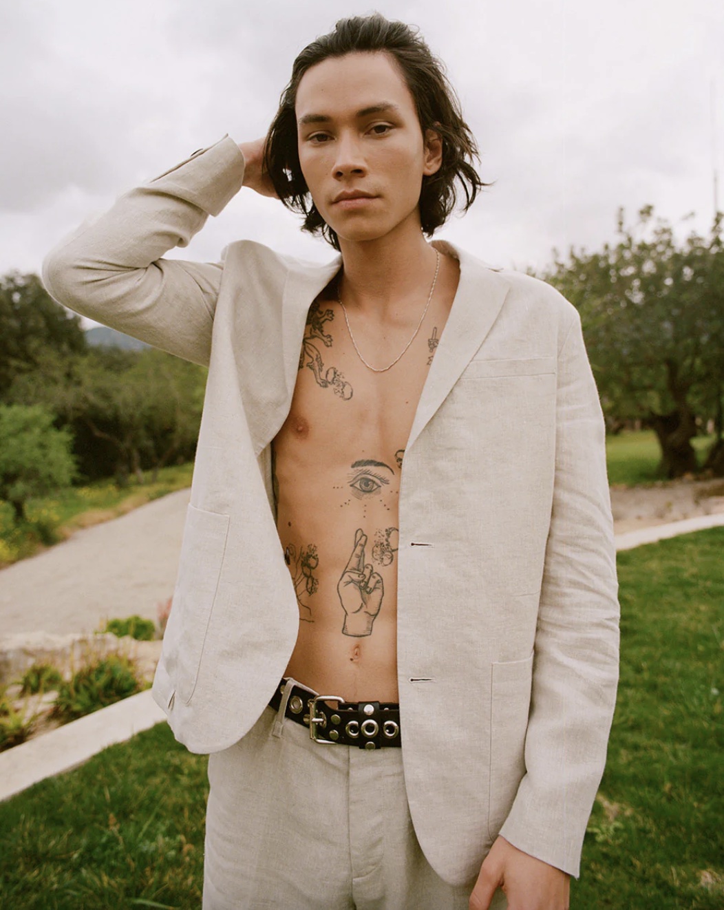Man wearing a linen blazer with no shirt underneath, standing outside with tattoos on chest