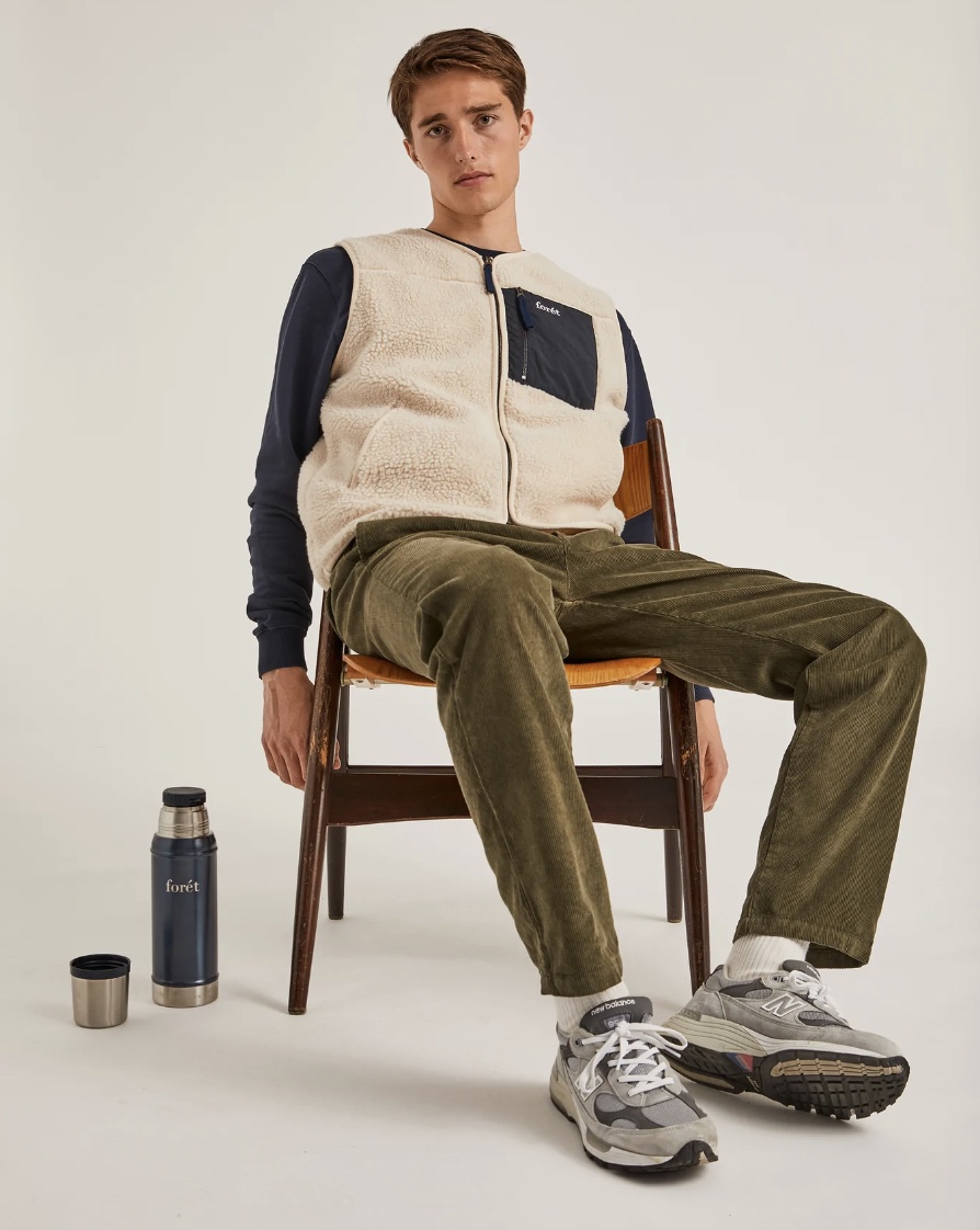 man slouching in a chair wearing green cords, runners and a vest with a thermos open on the ground