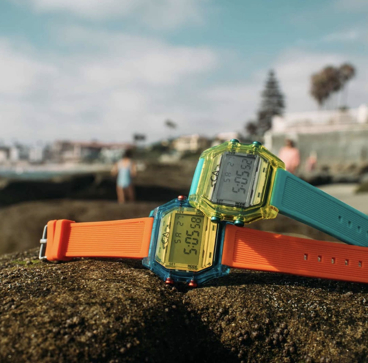 Two digital watches on rocks outside, one is orange and one is blue 