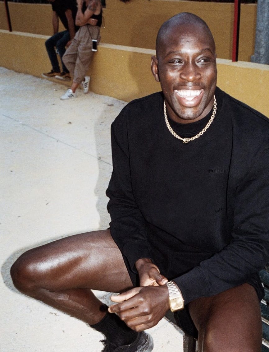 Man sitting, smiling wearing a black sweater, black shorts and a gold chain