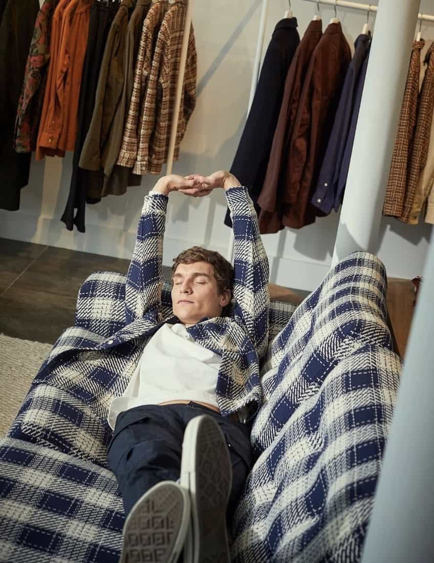 Man laying on a couch matching the same flannel he's wearing. Stretching his arms over his head .
