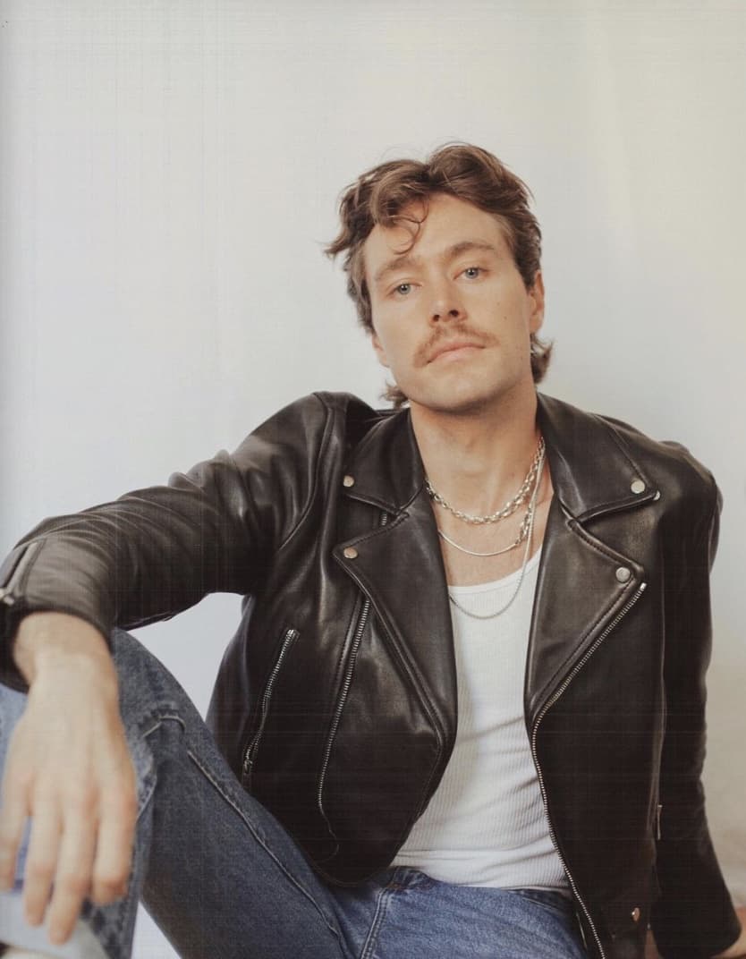 man sitting looking at the camera, wearing a leather jacket, tank top and jeans 