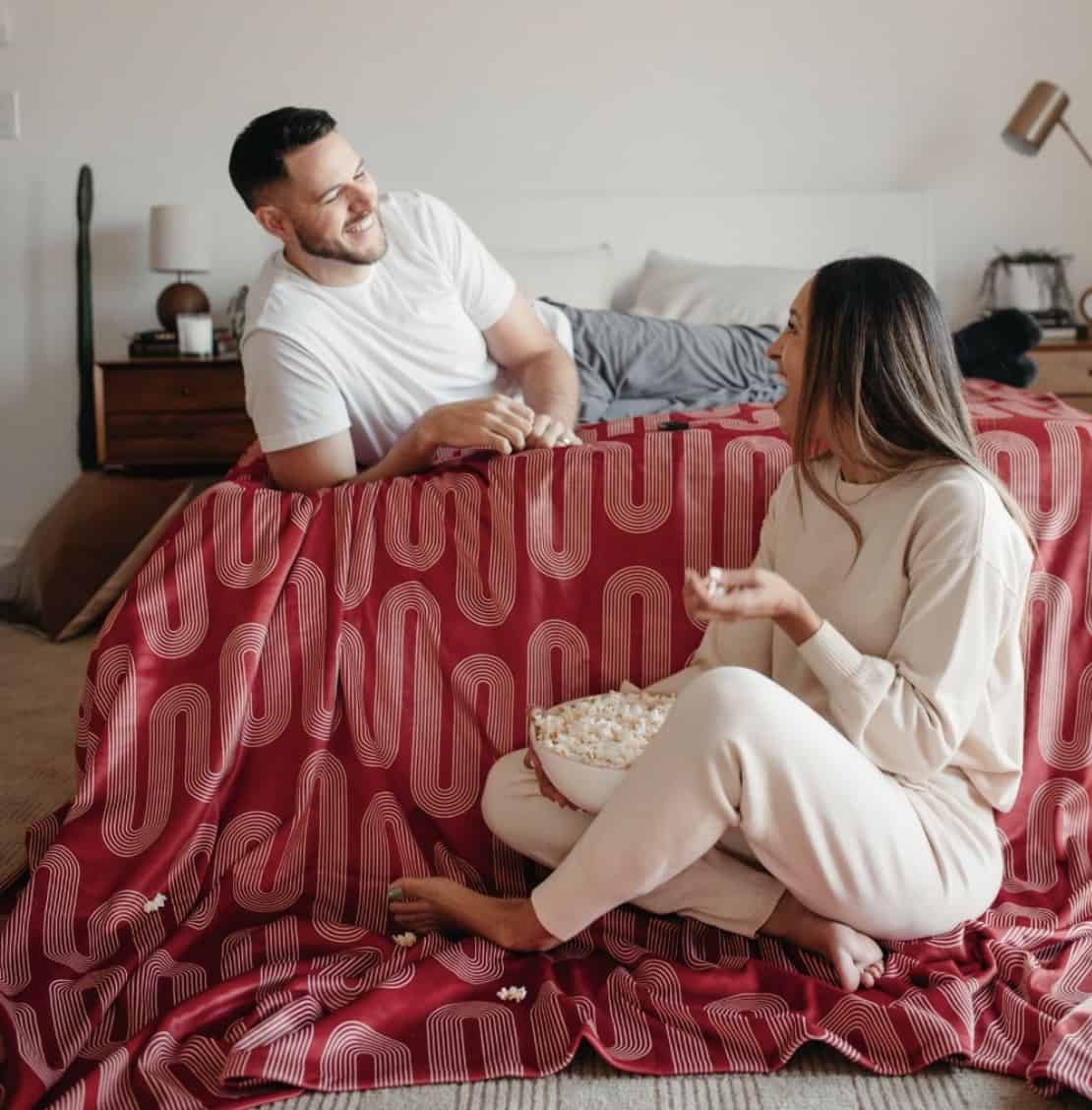 man laying on bed, women sitting on the ground eating popcorn, looking at each other